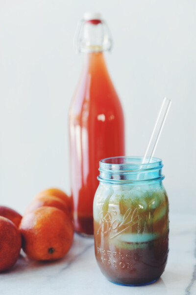 Homemade water kefir drink in a blue mason jar glass with ice and a glass straw next to a flip top glass bottle of water kefir with citrus fruit next to it.