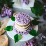 Angled shot of a mini 3-layer vegan lilac lemon cake with whipped coconut cream frosting on a white cake stand with lilac flowers and leaves around the base of the cake. The top of the cake is decorated with fresh lilac flowers. The background is styled with lilac flowers, tea dyed cheesecloth, a vintage lilac colored glass bottle, and a flower guide opened to a page on lilacs.