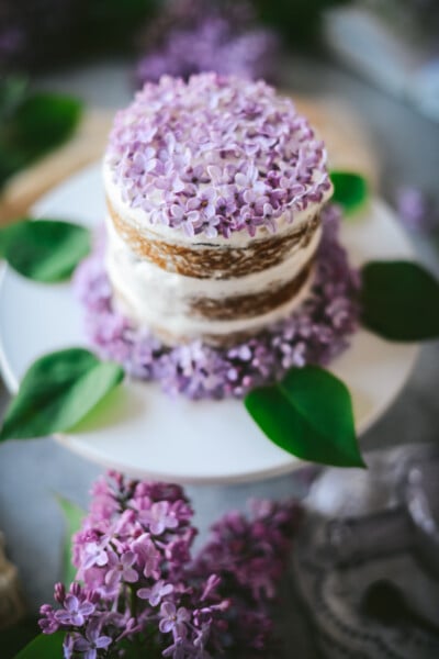 Closeup angled shot of a mini 3-layer vegan lilac lemon cake with whipped coconut cream frosting on a white cake stand with lilac flowers and leaves around the base of the cake. The top of the cake is decorated with fresh lilac flowers.