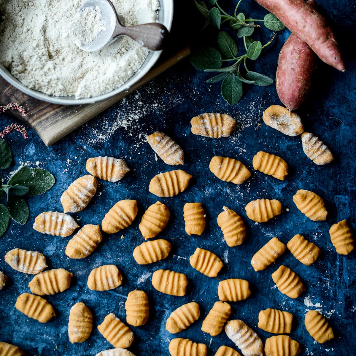 Uncooked vegan 3-ingredient sweet potato gnocchi shaped by hand on a blue background next to raw whole sweet potatoes, sage leaves, and a bowl of flour with a wooden scoop in it.