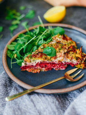 A slice of gluten free vegan tofu quiche with a potato crust on a blue plate with watercress salad and a brass fork.