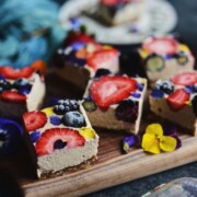 Raw vegan cheesecake bars topped with sliced strawberries, blackberries, blueberries and edible flowers on a wood board