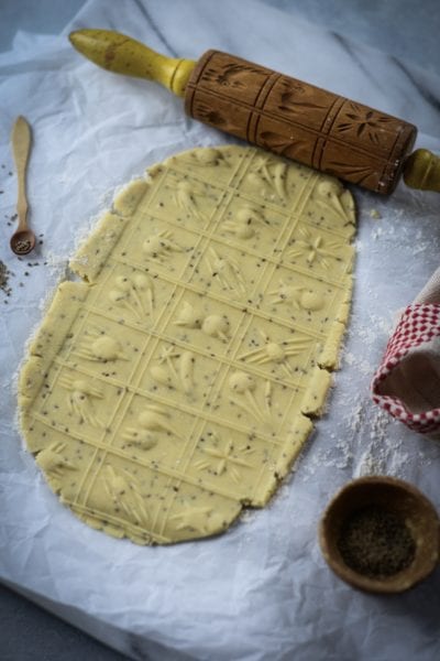Carved rolling pin on parchment with an embossed pattern on rolled-out cookie dough. A red and white cloth, a wooden spoon with spices, and a small bowl of black pepper are nearby.