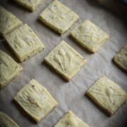 Square shortbread cookies with flecks of black sesame seeds are placed evenly on a parchment-lined baking sheet. The cookies are light golden brown, showcasing a textured surface. The background is slightly blurred.