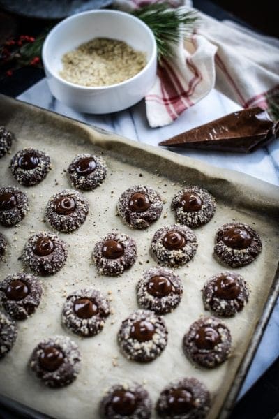A baking tray lined with parchment paper holds rows of chocolate thumbprint cookies filled with chocolate ganache and sprinkled with chopped nuts. In the background, a bowl of crushed nuts and a piping bag are visible.