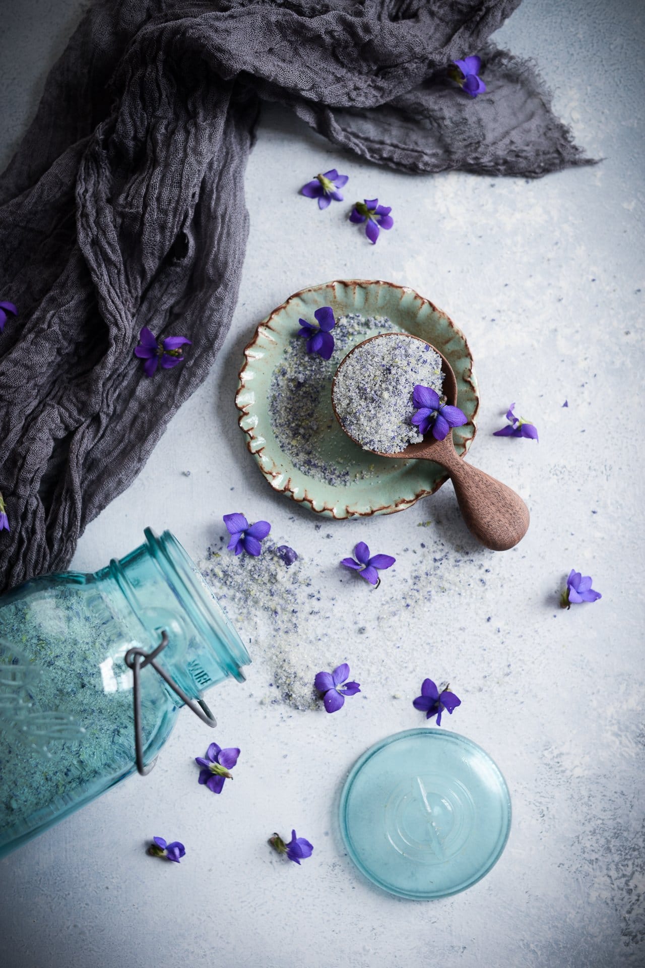 Top down view of violet sugar in a wooden scoop on a small green ceramic plate and a vintage blue glass mason jar on its side with violet sugar poured out of its mouth on a white background with violet flowers.