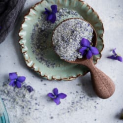 Top down closeup view of violet sugar in a wooden scoop on a small green ceramic plate on a white background with violet flowers.
