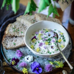 Vegan cashew creem cheese palted in a bowl and garnished with chive blossoms, chopped chives and edible flowers. Slices of bread are fanned out around the bowl.