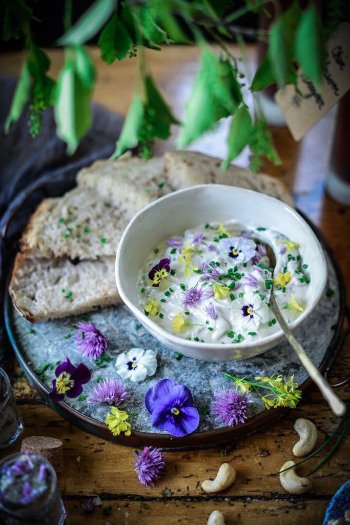 Vegan cashew creem cheese palted in a bowl and garnished with chive blossoms, chopped chives and edible flowers. Slices of bread are fanned out around the bowl.