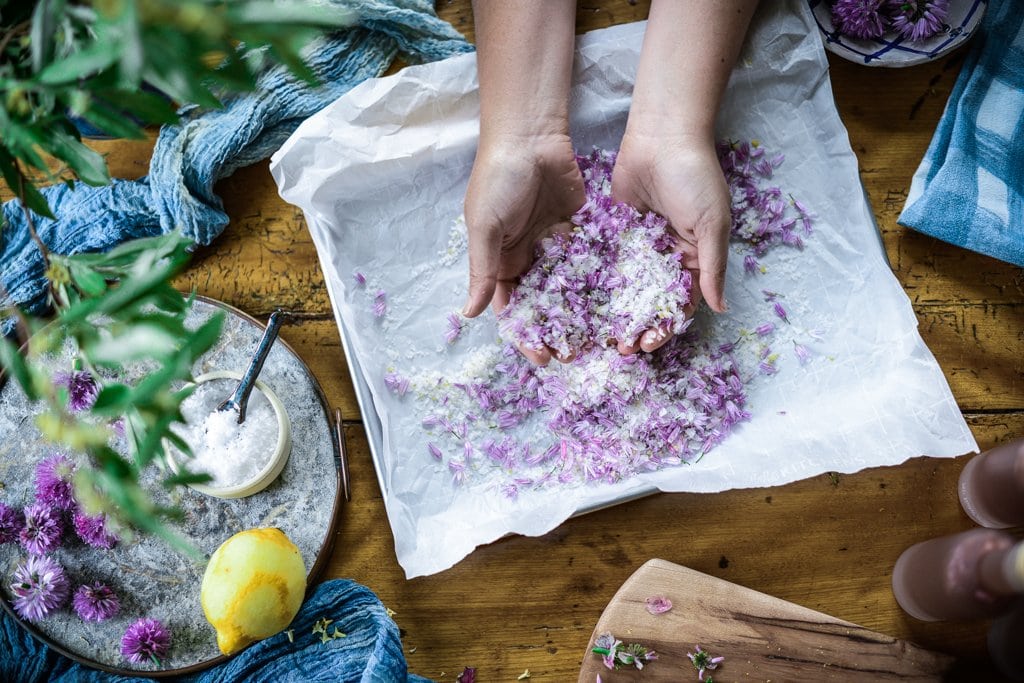 top down view of Kaity's hands mixing flaking sea salt, lemon zest and chive blossoms together on a parchment paper lined sheet pan.