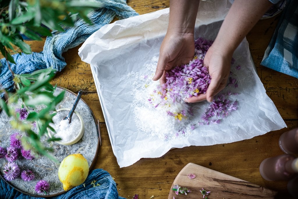 top down view of Kaity's hands mixing flaking sea salt, lemon zest and chive blossoms together on a parchment paper lined sheet pan.
