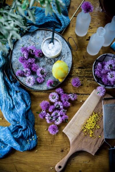 A rustic wooden table features purple chive blossoms perfect for flowering chive recipes, a lemon with grated zest on a cutting board, bowl of salt, blue cloth, and small glass bottles—all in a vibrant, colorful scene.