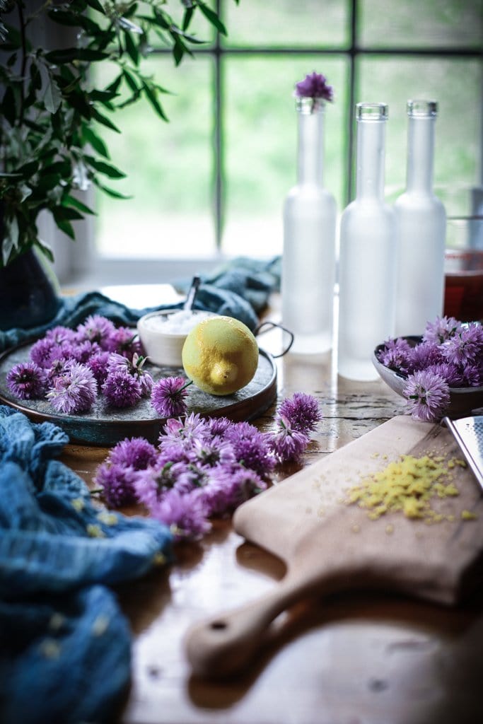 fresh picked chive blossoms are piled on a table. Grated lemon zest is on a small cutting board and the lemon sits nearby next to a dish of flaky salt. Three empty bottle sit it on the table.