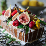A loaf cake with white icing, topped with fresh figs, raspberries, edible flowers, and green leaves, sits on a wire rack—an artful creation by a Nantucket private chef—with lemons blurred in the background.