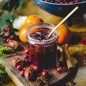 Maple spiced whole berry cranberry sauce in a glass jelly jar with a brass spoon in it surrounded by fresh cranberries, oranges and whole spices.