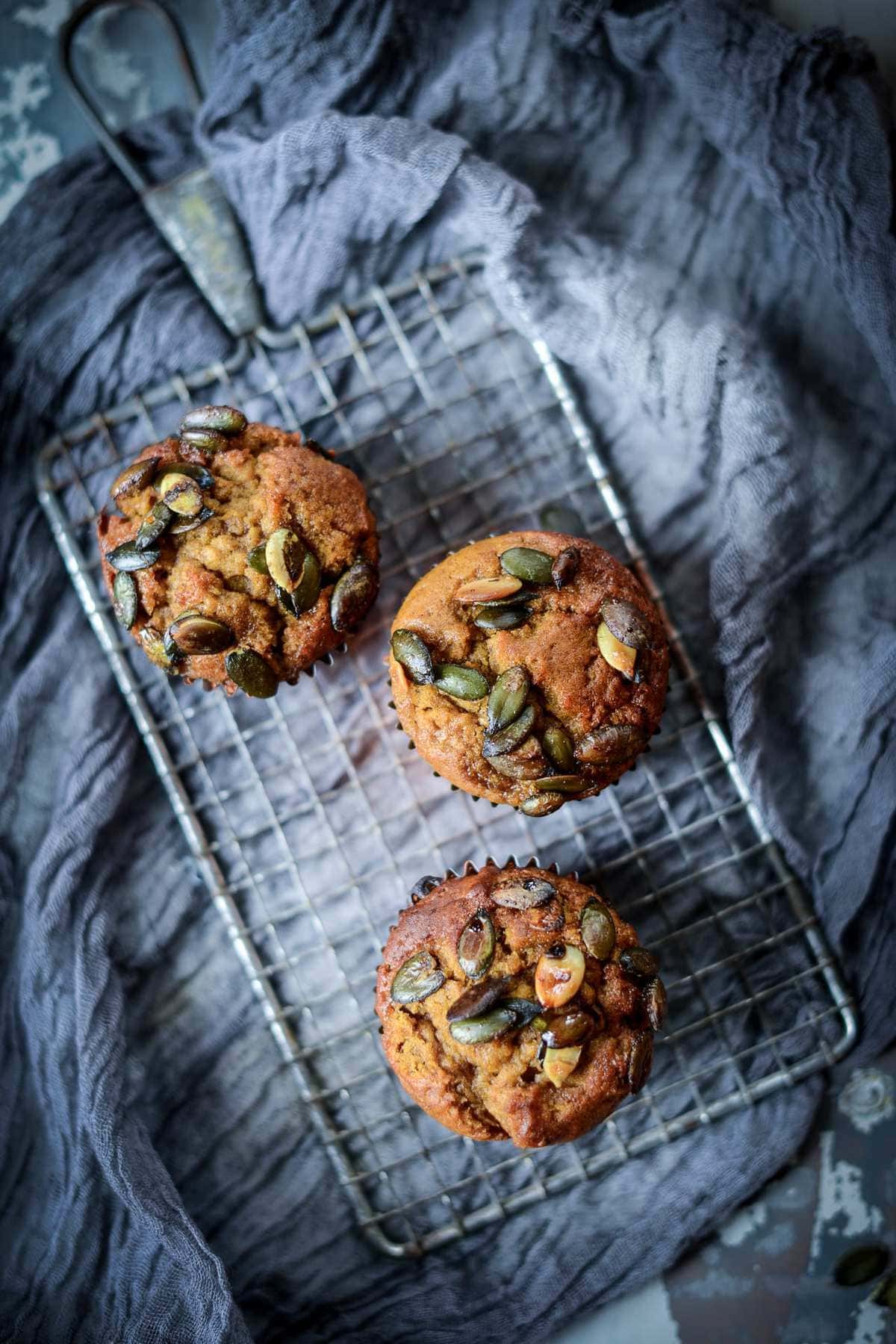 Three vegan pumpkin muffins topped with maple pumpkin seeds on a cooling rack over a grey cloth.