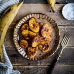 fried sweet plantains on a brown plate with a ripe yellow plantain and fork next to the plate.