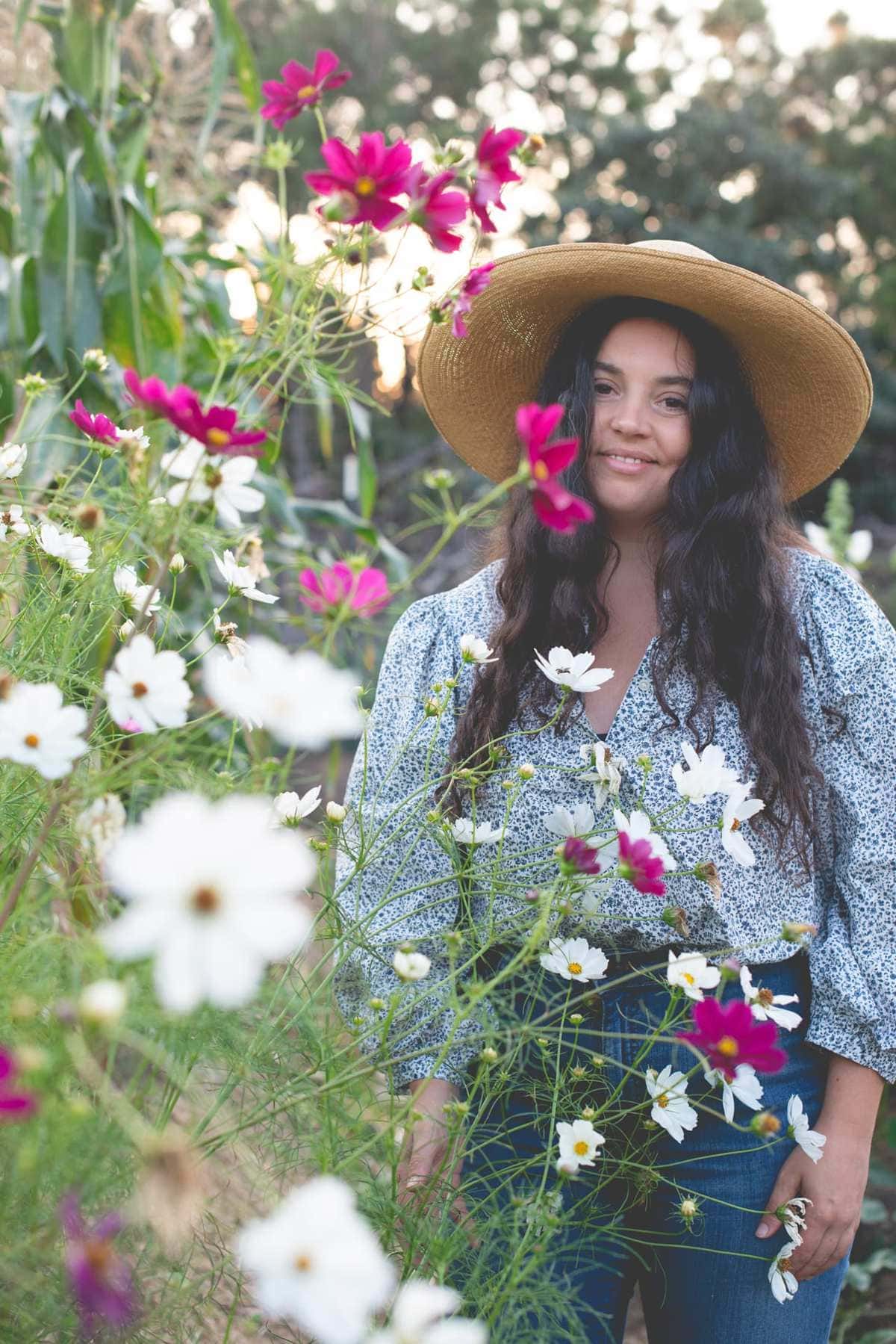 Kaity Farrell, owner of Fare Isle, wears a wide-brimmed straw hat and floral blouse as she stands among blooming white and pink wildflowers, smiling softly. Trees and sunlight are visible in the blurred background.