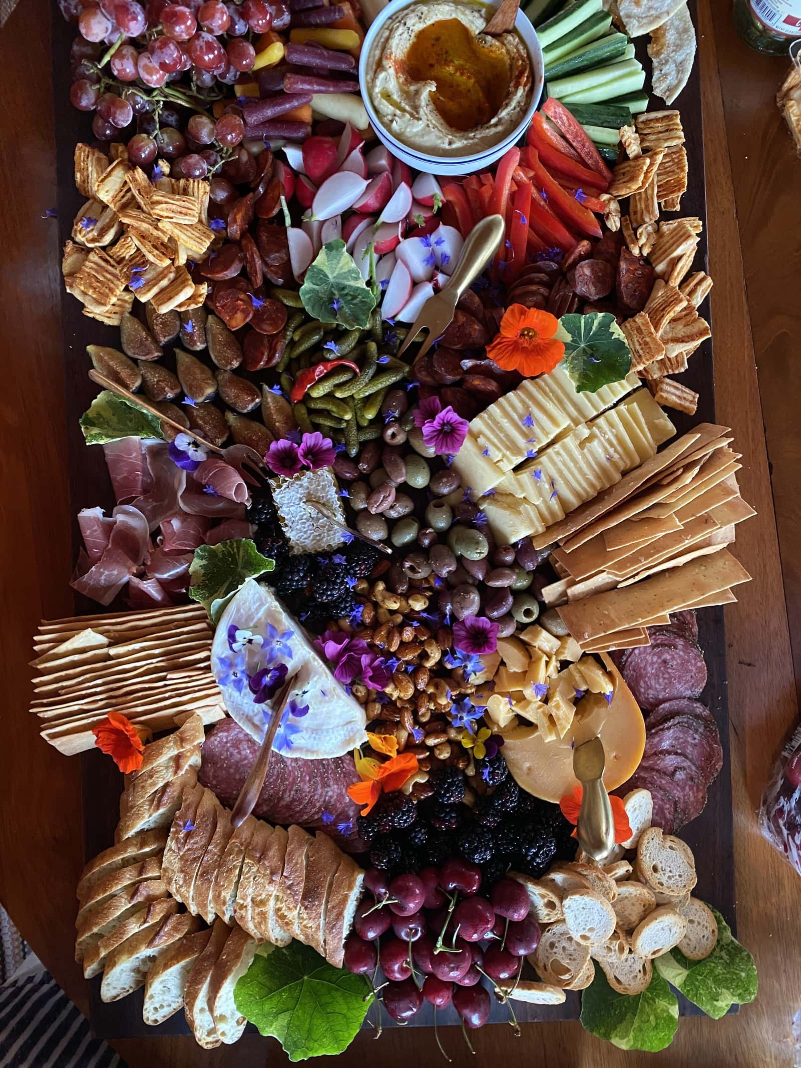 A large, colorful charcuterie board by Nantucket catering, filled with crackers, cheeses, cured meats, grapes, olives, nuts, fresh vegetables, berries, edible flowers, and a bowl of dip in the top center.