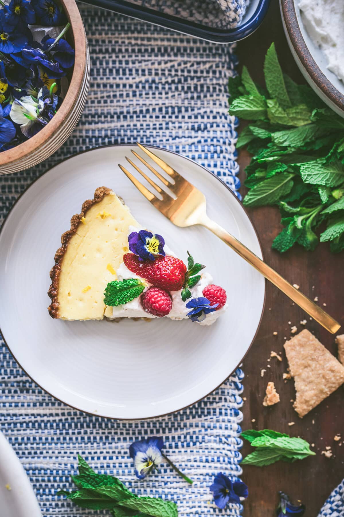 A slice of ricotta pie topped with whipped cream, raspberries, mint leaves, and edible purple flowers on a white plate with a gold fork, surrounded by fresh mint, flowers, and a woven blue placemat.