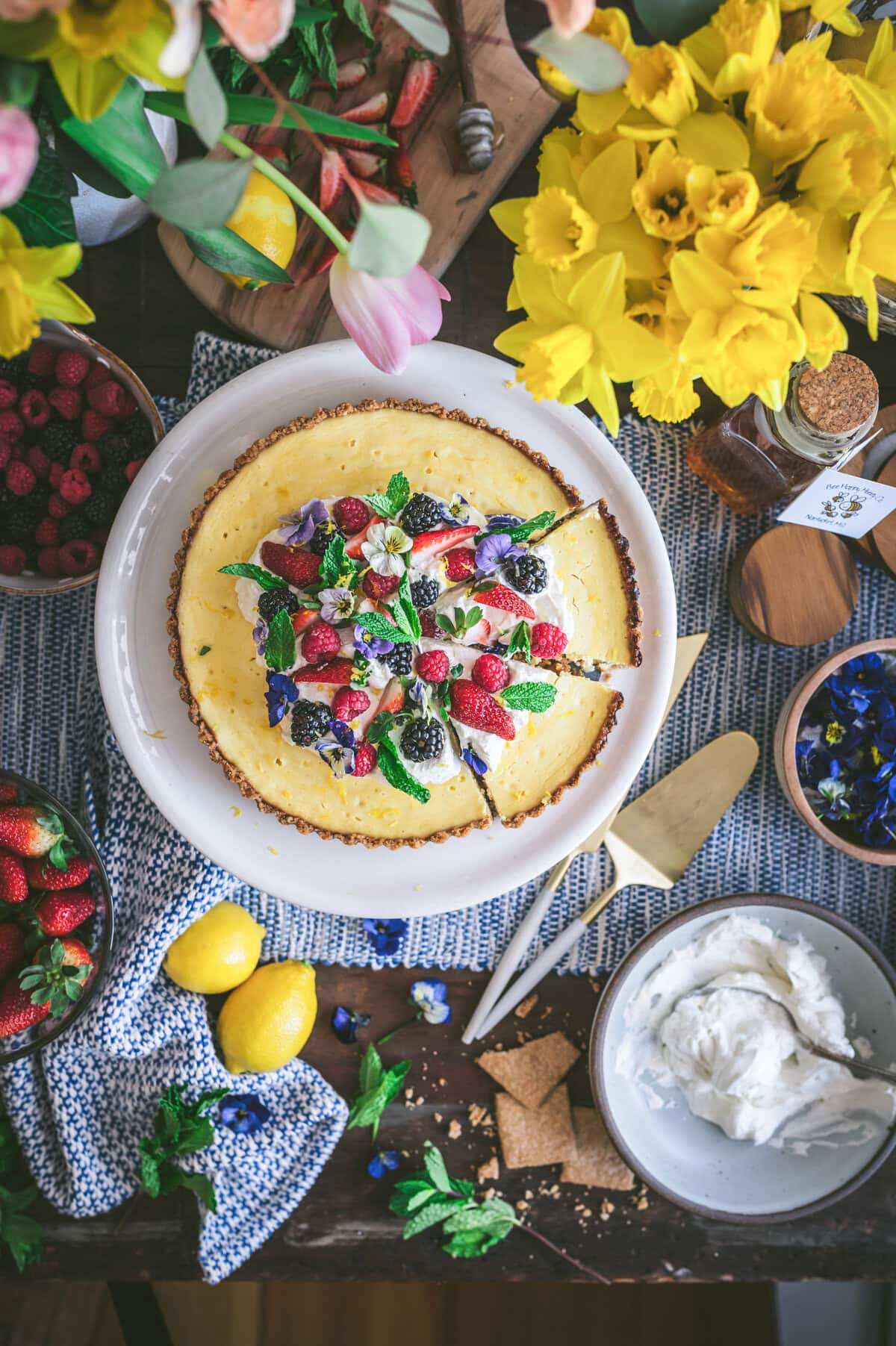 A classic ricotta pie is topped with assorted fresh berries, mint leaves, and edible flowers. It sits on a table surrounded by lemons, strawberries, whipped cream, graham crackers, and a bouquet of yellow flowers.