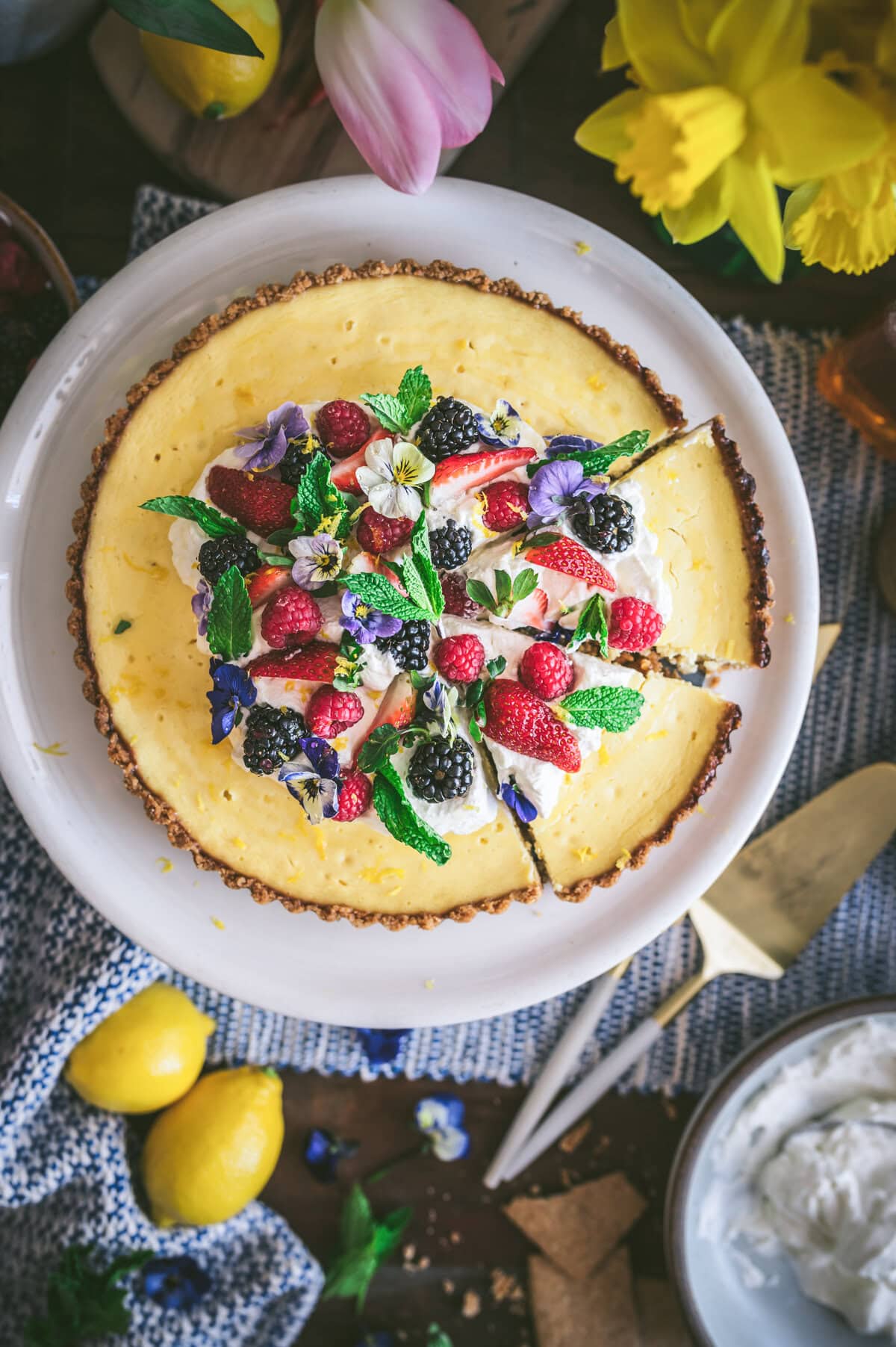 A ricotta pie cheesecake topped with strawberries, blackberries, raspberries, mint leaves, and edible flowers sits on a white platter. Two slices are cut. Lemons, whipped cream, and yellow flowers surround it.