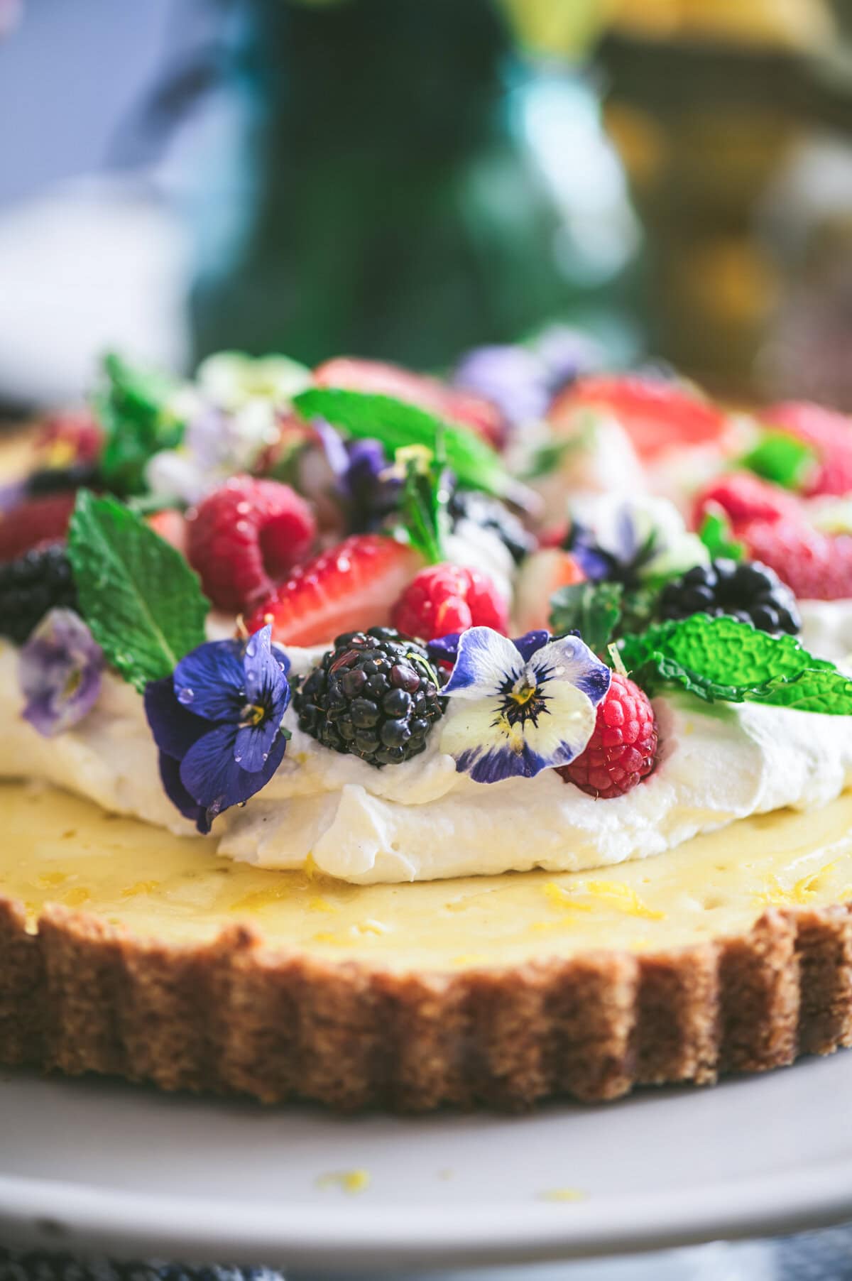 A close-up of a ricotta pie topped with whipped cream, fresh berries, mint leaves, and purple edible flowers, displayed on a plate with a soft-focus background.