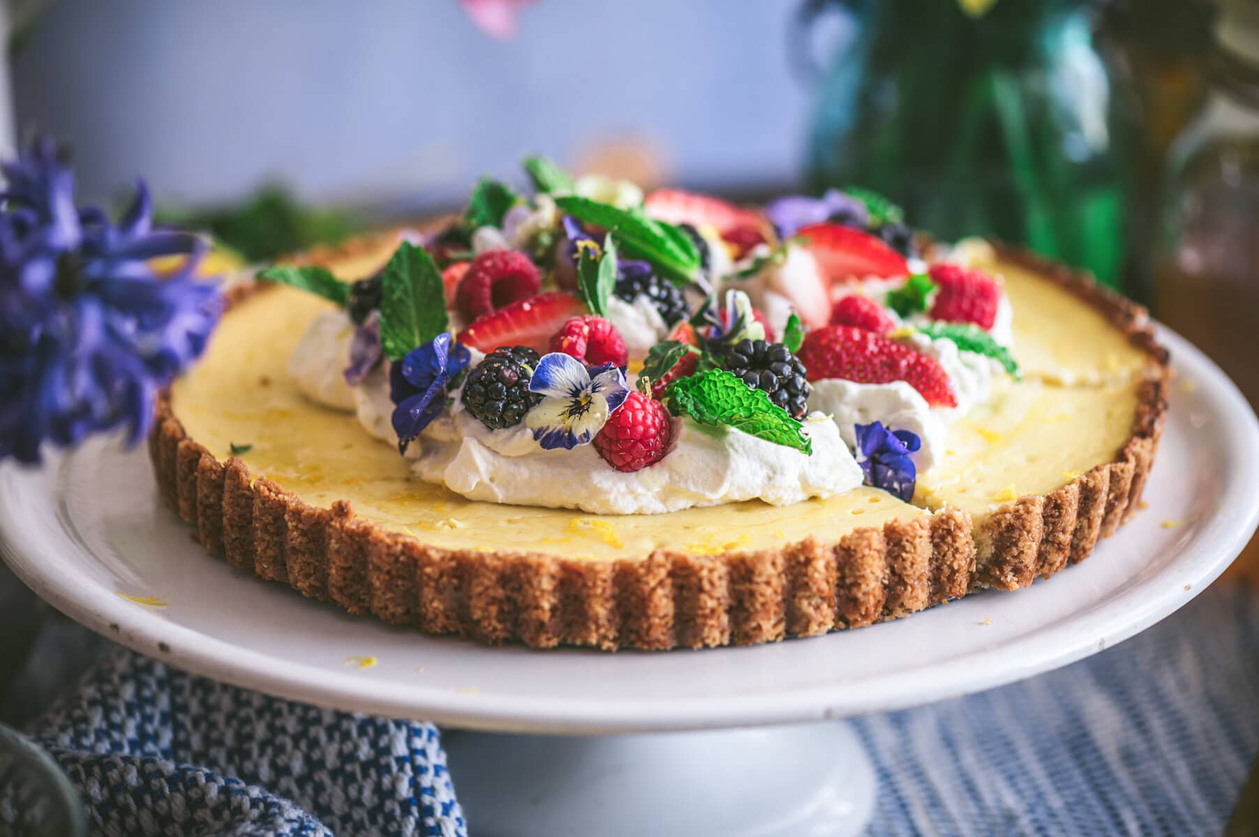 A ricotta pie on a white cake stand topped with whipped cream, fresh raspberries, blackberries, strawberries, mint leaves, and edible flowers, set on a patterned cloth with blurred flowers in the background.