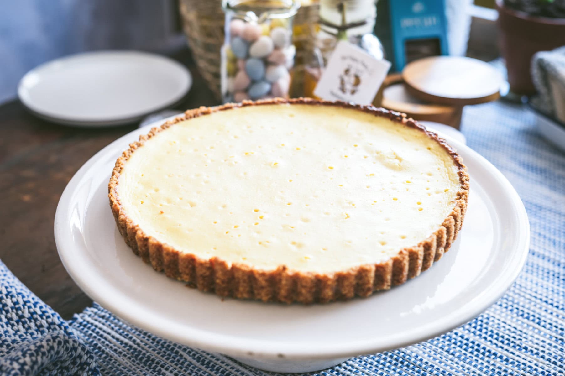 A ricotta pie with a graham cracker crust sits on a white cake stand, with jars of pastel candies and tableware softly blurred in the background.