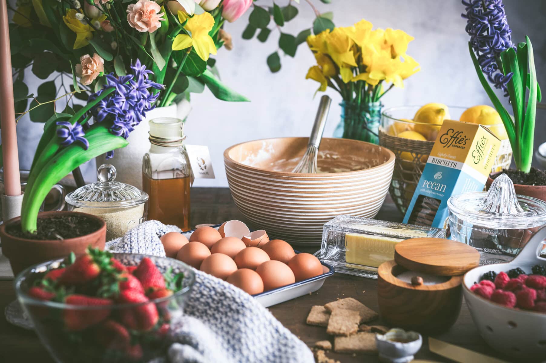 A kitchen table filled with baking ingredients—eggs, butter, biscuits, strawberries, honey—and the makings of a ricotta pie sits among colorful spring flowers and potted plants, creating a bright, inviting scene.