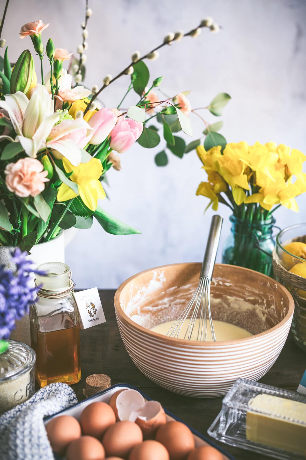 A mixing bowl with batter for ricotta pie and a whisk, surrounded by eggs, butter, honey, and fresh flowers, including tulips and daffodils, on a kitchen counter.