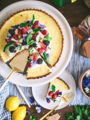 A ricotta pie topped with fresh strawberries, blackberries, mint, and edible flowers sits on a cake stand. One slice is served on a plate below, surrounded by lemons, oat cakes, and scattered flowers.