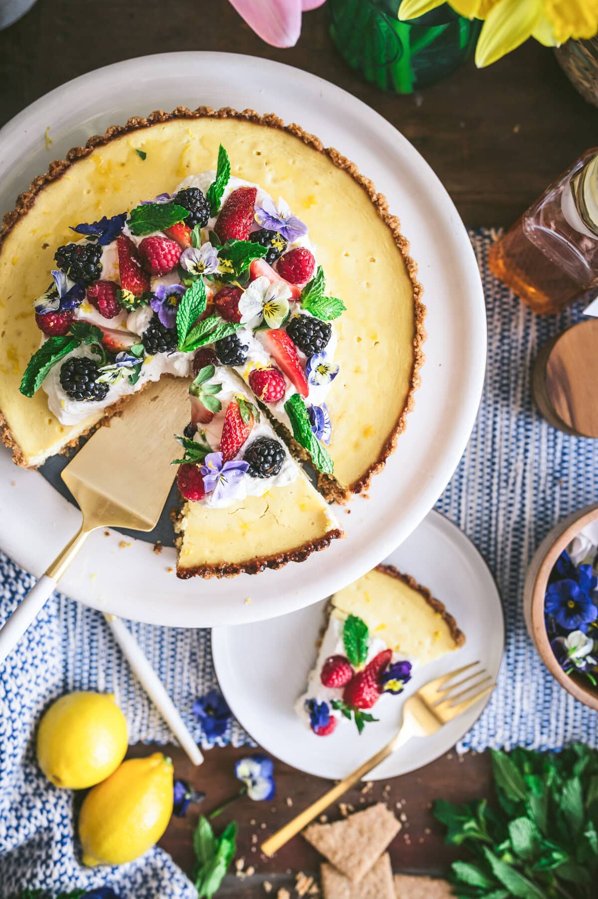A ricotta pie topped with fresh strawberries, blackberries, mint, and edible flowers sits on a cake stand. One slice is served on a plate below, surrounded by lemons, oat cakes, and scattered flowers.