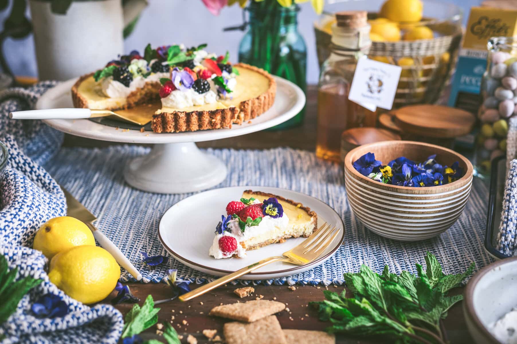 A table set with a ricotta pie topped with whipped cream and edible flowers, a slice on a plate with a gold fork, lemons, herbs, honey, biscuits, and a blue patterned cloth.