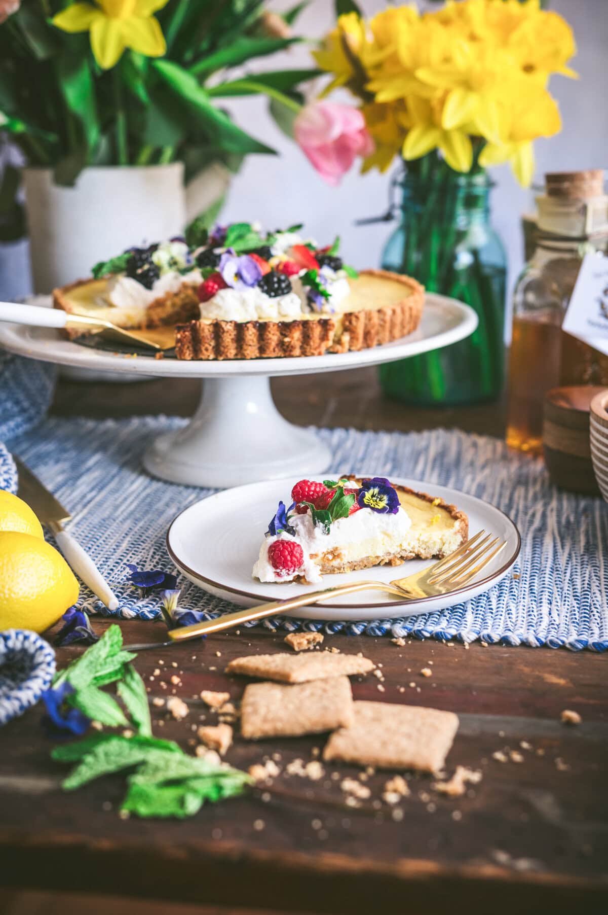 A slice of fruit and flower-topped ricotta pie on a plate with a fork sits in front of the whole tart on a cake stand, surrounded by spring flowers, lemons, and graham crackers on a wooden table.