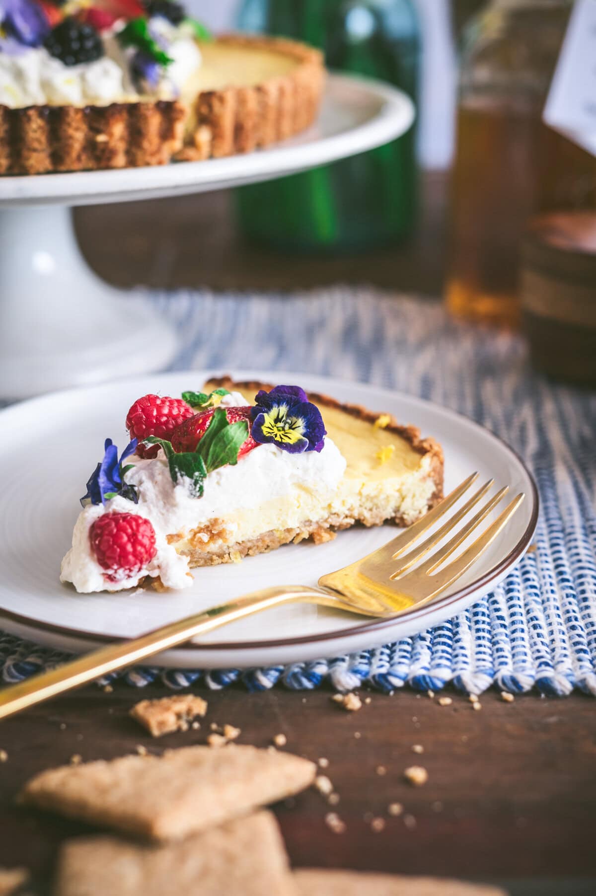 A slice of ricotta pie topped with whipped cream, fresh raspberries, mint leaves, and edible flowers sits on a white plate with a gold fork. The whole tart is in the background on a cake stand.