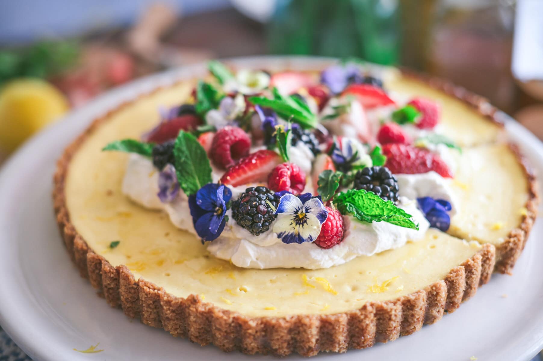 A ricotta pie-inspired cheesecake tart topped with whipped cream, fresh strawberries, blackberries, raspberries, mint leaves, and edible purple flowers, beautifully displayed on a white plate.