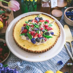 A ricotta pie cheesecake topped with berries, mint leaves, and edible flowers sits on a white plate. Surrounding it are lemons, flowers in vases, honey, nuts, and fruits on a textured blue cloth.