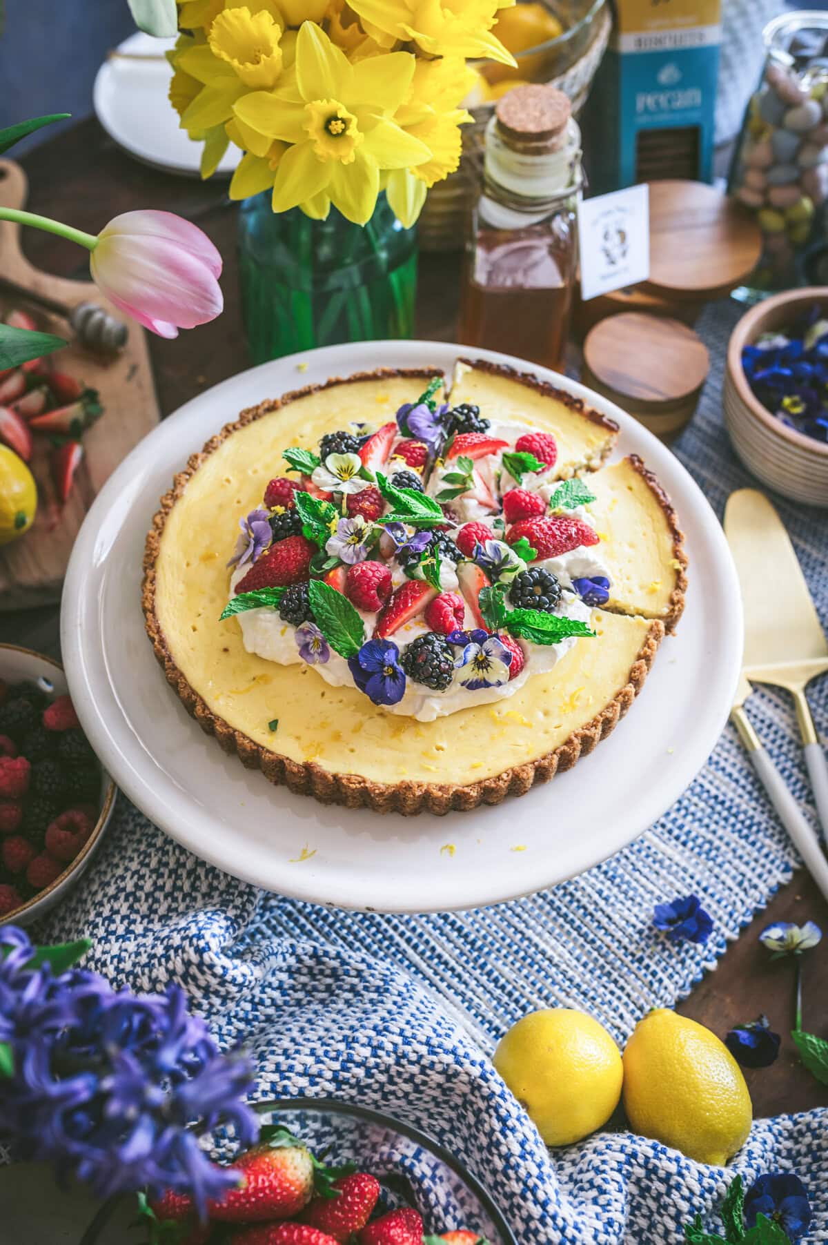 A ricotta pie cheesecake topped with berries, mint leaves, and edible flowers sits on a white plate. Surrounding it are lemons, flowers in vases, honey, nuts, and fruits on a textured blue cloth.