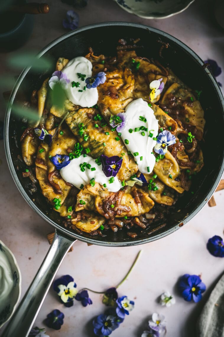 Finished dish of edible flower pierogi are plated in a skillet with sauteed onions and topped with sour cream, chives and edible flowers.
