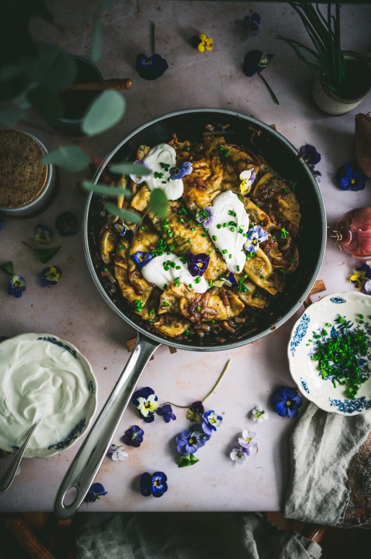 Finished dish of edible flower pierogi are plated in a skillet with sauteed onions and topped with sour cream, chives and edible flowers.