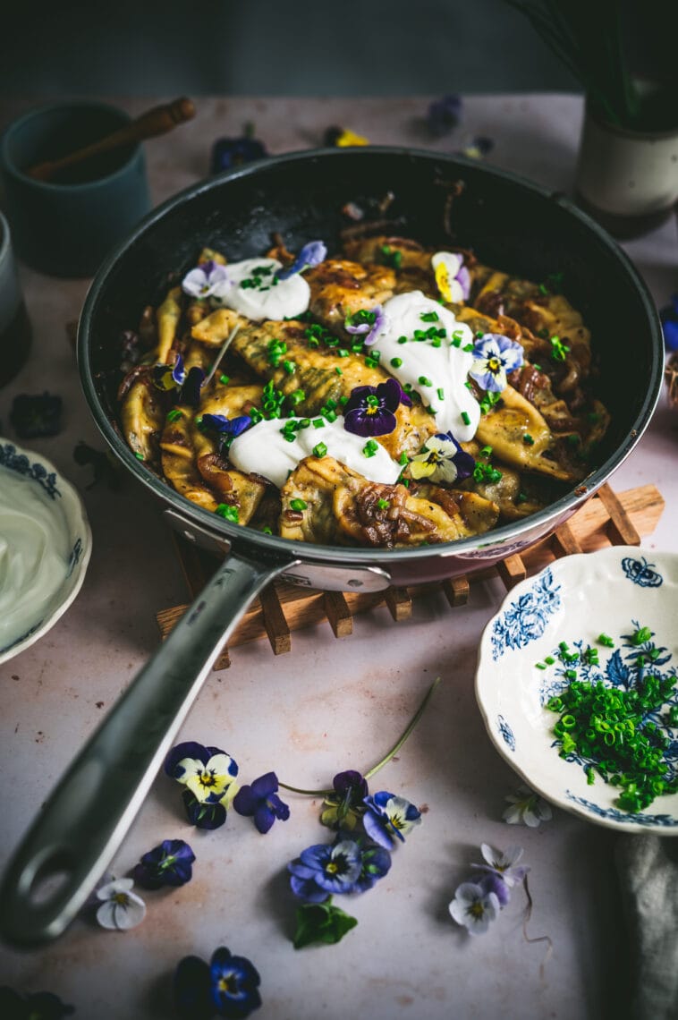Finished dish of edible flower pierogi are plated in a skillet with sauteed onions and topped with sour cream, chives and edible flowers.