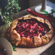 A rustic strawberry rhubarb galette with a golden, folded crust sits on a wooden cutting board, surrounded by flowers, a bowl of fruit, and a candle in the background.