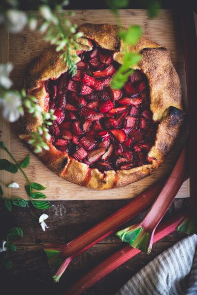 A rustic strawberry rhubarb galette with a golden, flaky crust sits on a wooden board, surrounded by fresh rhubarb stalks and green leaves, with flowers and a striped cloth nearby.