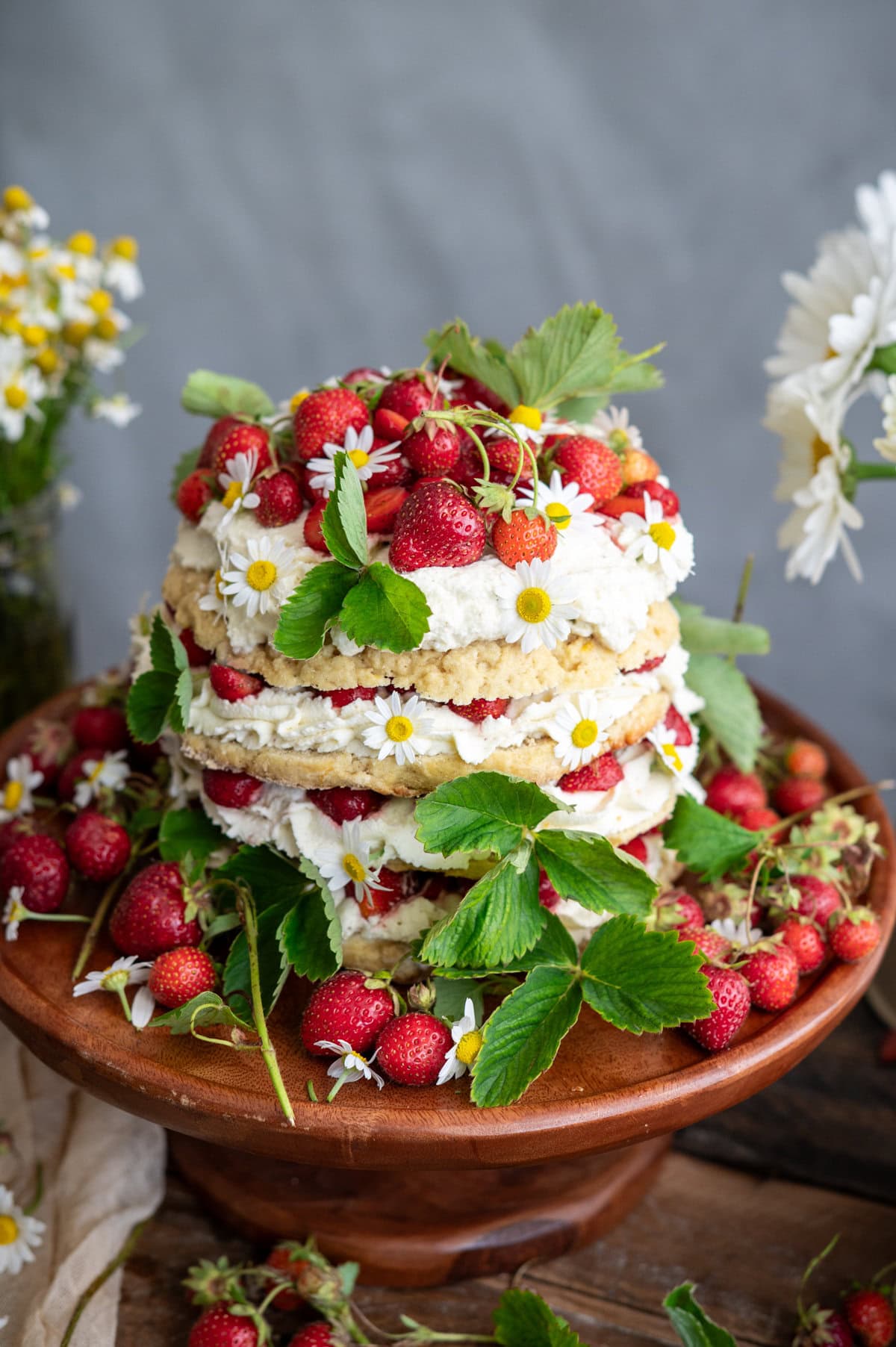 A layered strawberry shortcake cake decorated with fresh strawberries, whipped cream, green leaves, and small daisies sits on a wooden cake stand, surrounded by more strawberries and flowers.