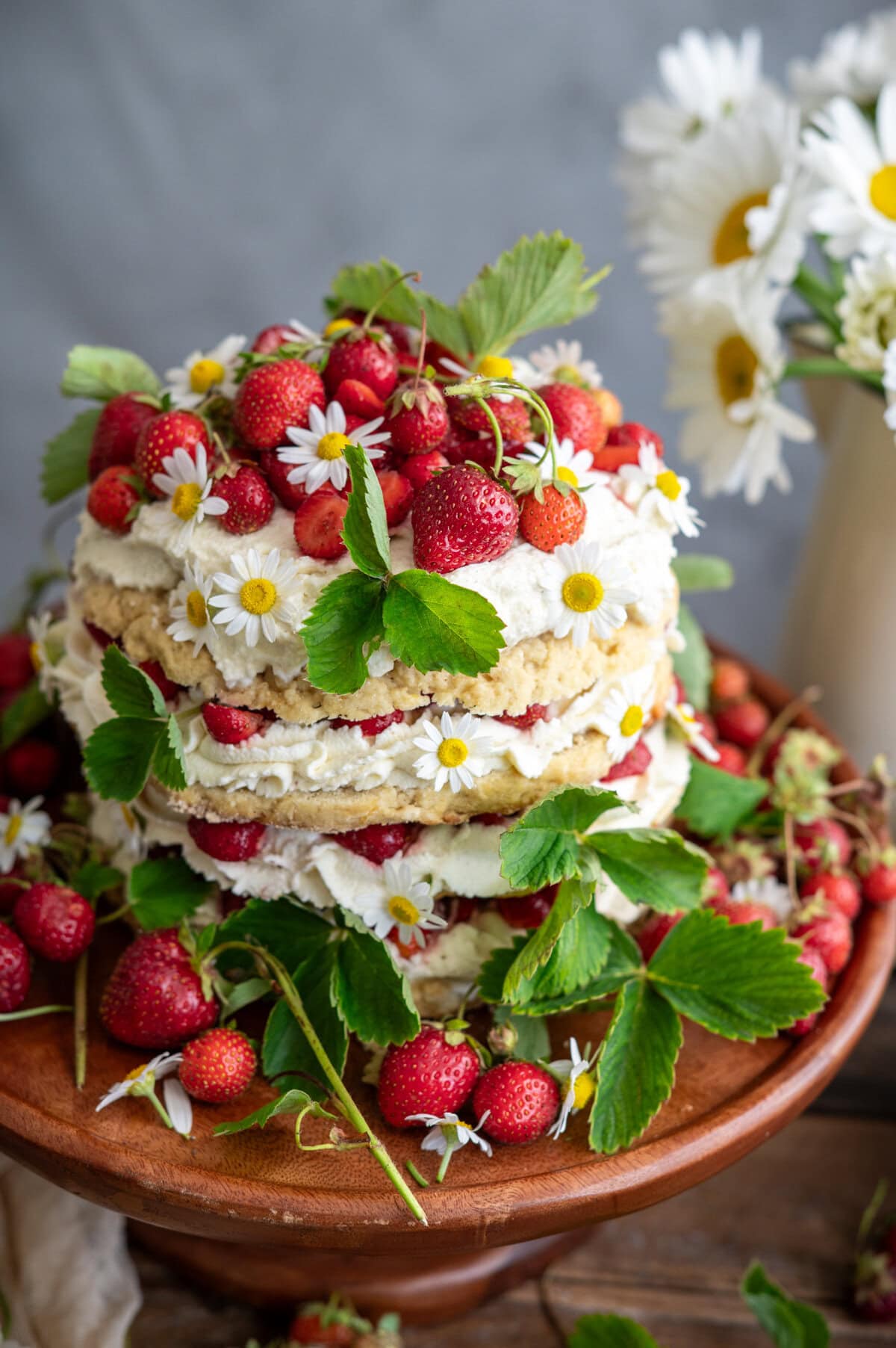 A rustic strawberry shortcake cake with cream and fresh strawberries, decorated with strawberry leaves and small white daisies, sits on a wooden cake stand. More strawberries and flowers are scattered around, with a vase of daisies in the background.