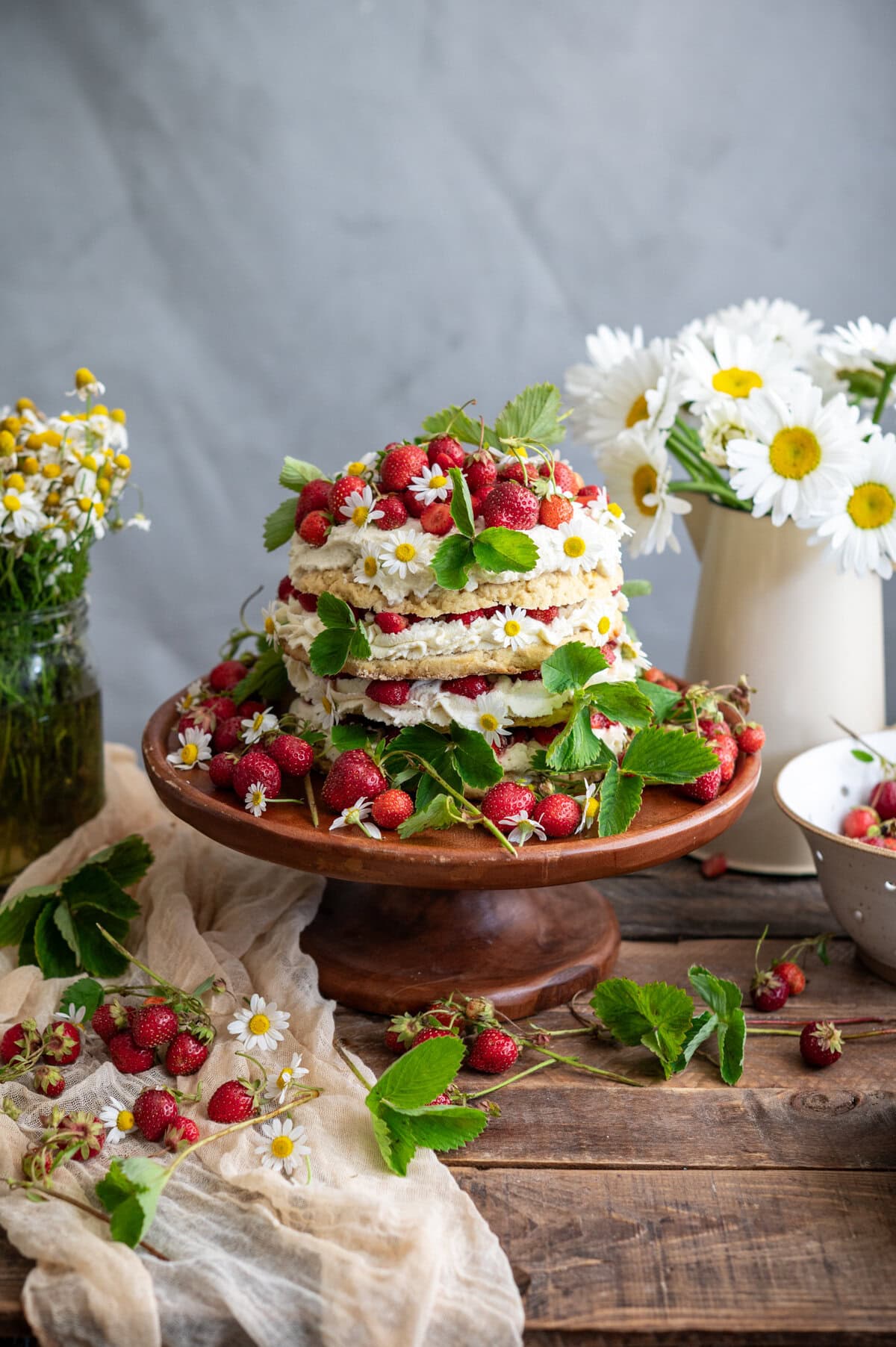 A rustic strawberry shortcake cake decorated with fresh strawberries and green leaves sits on a wooden cake stand. Wildflowers and daisies surround the cake, with more strawberries scattered across the wooden table.