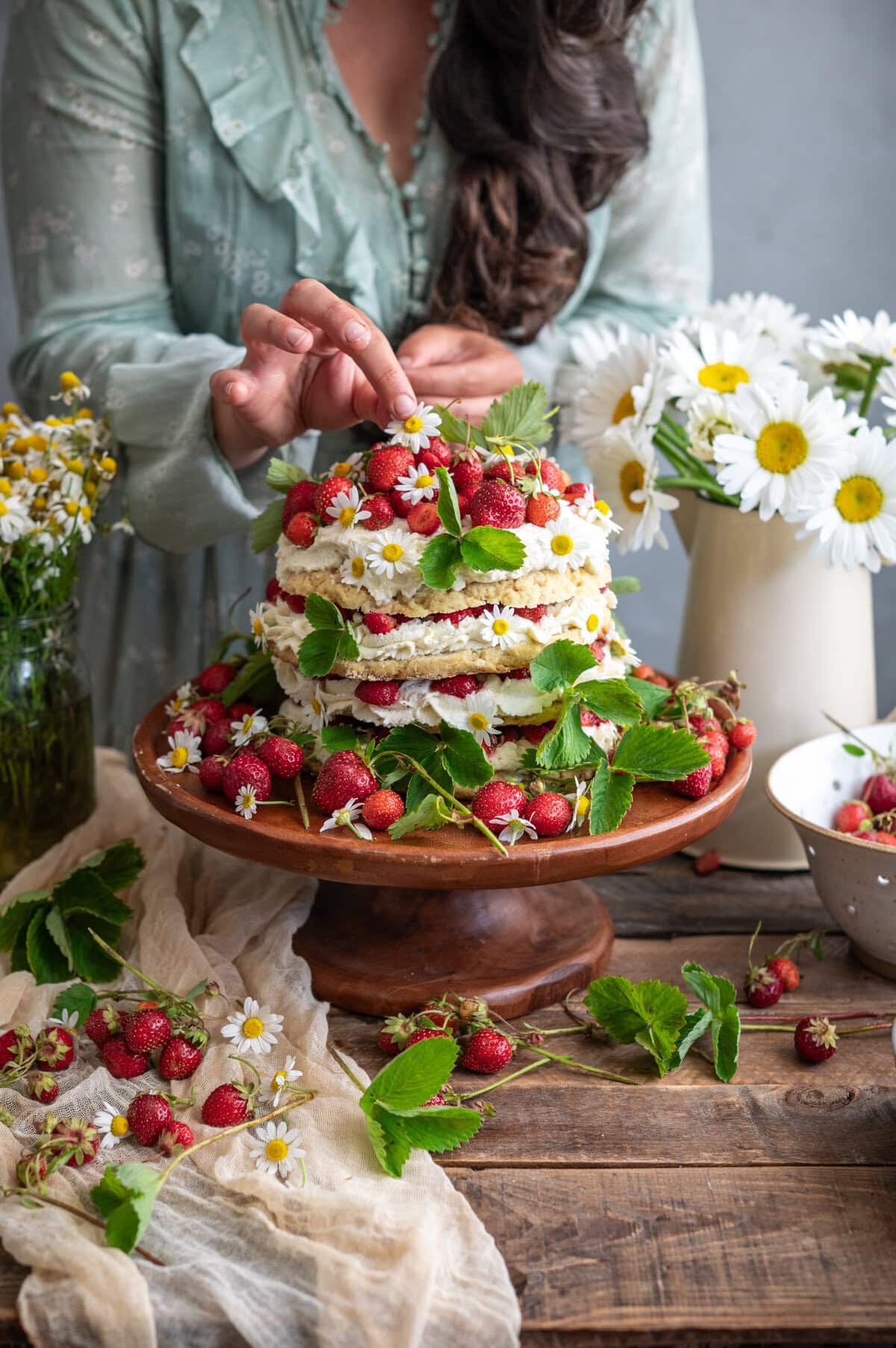 A woman in a light green dress decorates a strawberry shortcake cake with strawberries and leaves on a wooden cake stand, surrounded by fresh strawberries, leaves, and vases of daisies on a rustic wooden table.
