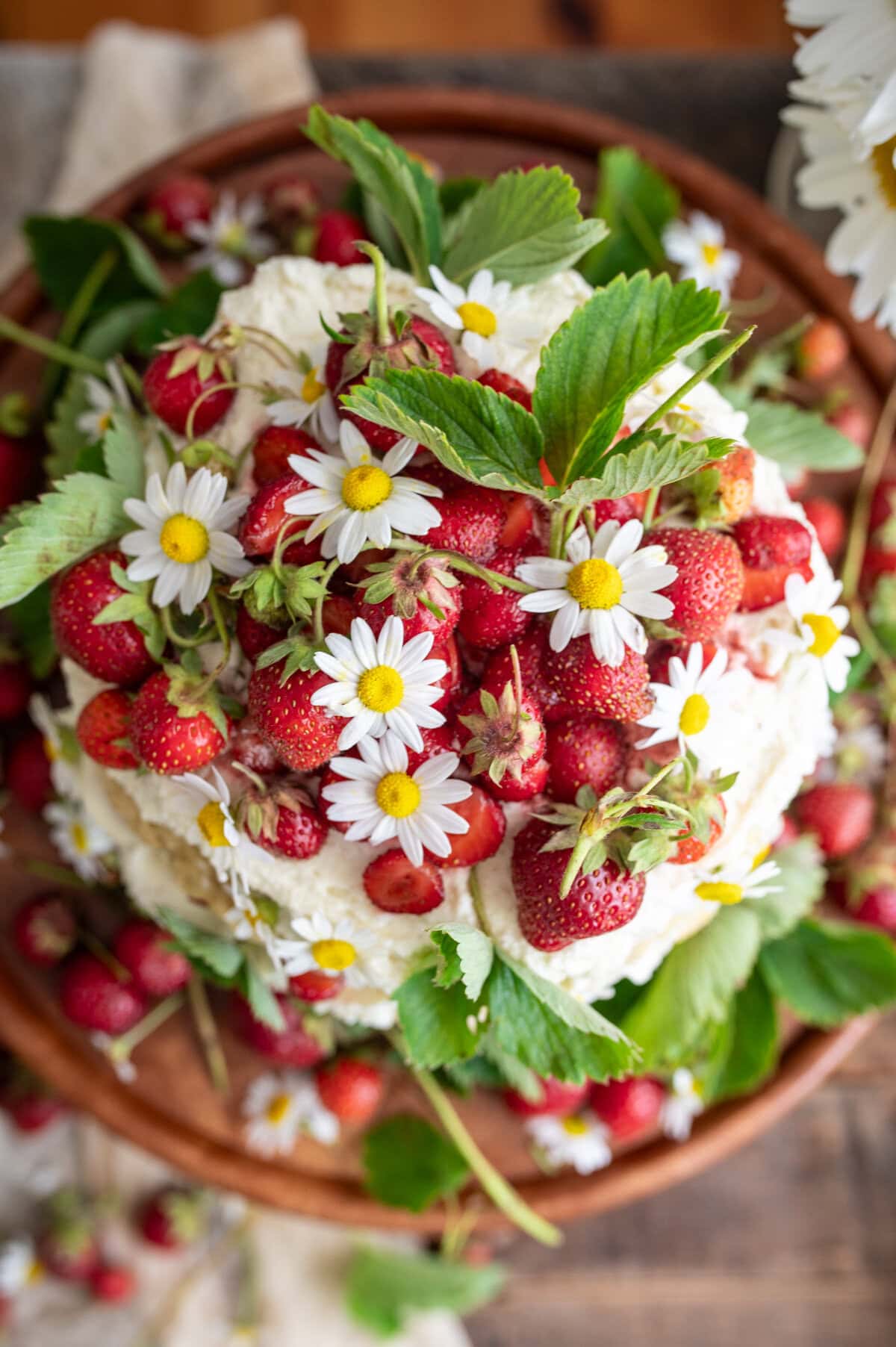 A delightful strawberry shortcake cake topped with fresh strawberries, whipped cream, green leaves, and white daisies, presented on a wooden plate with extra strawberries and flowers artfully scattered around.