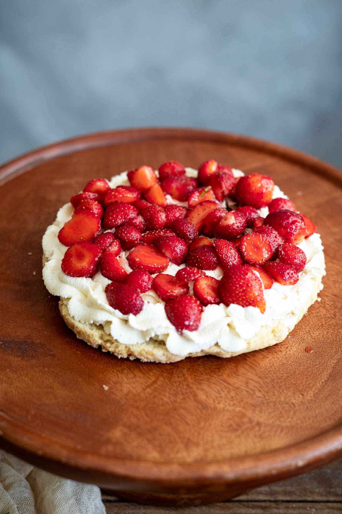 A strawberry shortcake cake topped with whipped cream and sliced fresh strawberries sits on a wooden cake stand against a neutral background.